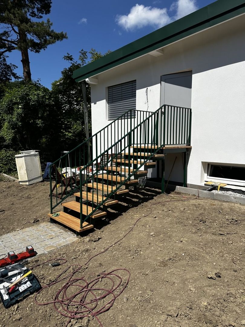 Eine Podesttreppe aus Holz mit einem schwarzen Metallgeländer führ zu einer Haustür eines weißen Hauses