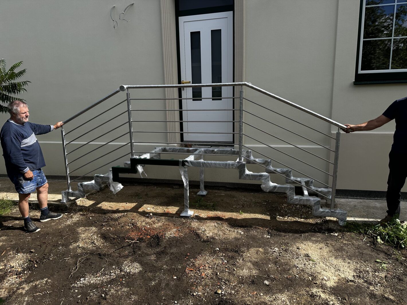 Zwei Mitarbeiter arbeiten am Aufbau einer Zweiholmtreppe mit Metallgeländer vor einem Hauseingang